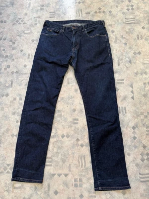 Mörkblåa raka Emporio Armani jeans  - Snygga Emporio Armani mörkblå jeans med rak passform och klassisk femficksdesign. Jeansen har en enkel look utan slitningar och är tillverkade i ett robust denimtyg. Perfekta för dig som gillar stilrena och tidlösa plagg. Nypris 2300kr kvitto finns på mailen. Storlek 33/32
