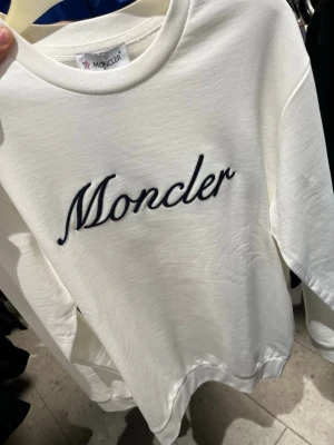 Vit sweatshirt från Moncler - Säljer en stilren vit sweatshirt från Moncler med broderad logga i svart på bröstet. Tröjan har rund halsringning, långa ärmar och ribbade muddar. Perfekt för dig som gillar exklusiva streetwear-plagg.