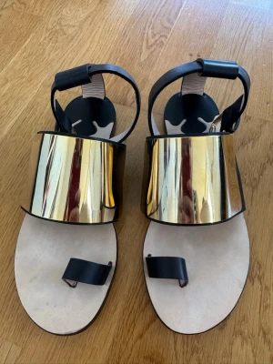 Svarta sandaler med gulddetalj strl 36 - Unika sandaler med bred guldfärgad metallpanel över foten och svarta läderremmar. Platt sula i beige och justerbar rem runt ankeln. Snygg och modern design som sticker ut och passar till många olika stilar.  Sandalerna är endast provade och aldrig använda utomhus. De är i mycket fint skick, men kan ha mindre märken från förvaring och provning. Storlek 36