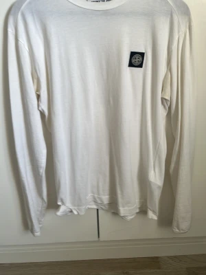 Vit långärmad T-shirt  från Stone Island - Vit långärmad T-shirt från Stone Island med klassisk rund hals och ikonisk logopatch på bröstet. Tillverkad i mjuk bomull för en bekväm känsla och clean look. Perfekt för dig som gillar stilrena plagg med diskreta detaljer.