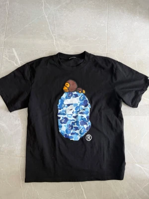 Svart BAPE t-shirt med blå kamotryck - Säljer en svart t-shirt från BAPE med ikoniskt blått kamotryck och en apa på bröstet. T-shirten har rund hals och korta ärmar. Perfekt för dig som gillar streetwear och vill sticka ut med en unik design.