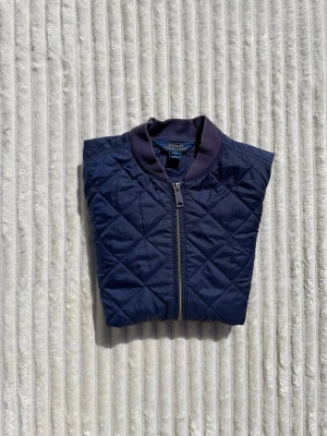 Marinblå quiltad bomberjacka Ralph Lauren - Snygg marinblå bomberjacka från Polo Ralph Lauren med quiltat mönster, ribbade muddar och krage. Jackan har dragkedja framtill och två fickor med tryckknappar. Perfekt för dig som vill ha en stilren och klassisk look.