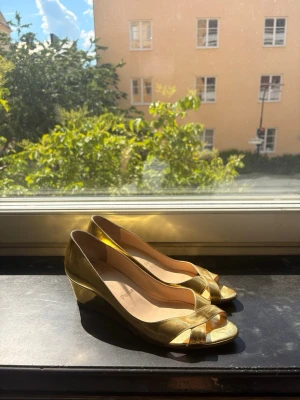 Guldiga pumps med öppen tå - Säljer ett par glansiga guldiga pumps med öppen tå och låg klack. Skorna har korsade remmar framtill och en klassisk siluett. Märkt med 'Paris' på innersulan. Perfekta för att sticka ut och ge en lyxig touch till din outfit.
