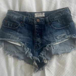 Blå jeansshorts med fransar - Säljer ett par blå jeansshorts från One Teaspoon med råa fransade kanter och slitna detaljer. Klassisk femficksmodell med knapp och dragkedja framtill. Perfekta för sommaren och ger en cool, avslappnad look.