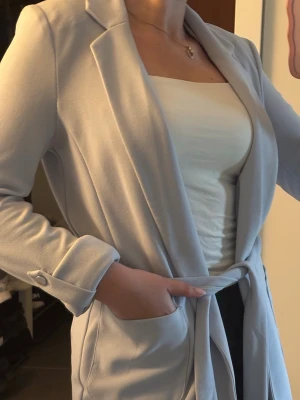 Ljusblå kappa från primark - Snygg ljusblå kappa från primark med klassisk krage, knytskärp i midjan och stora fickor framtill. Kappan har långa ärmar med knappdetalj vid mudden och är tillverkad i ett mjukt tyg som ger en stilren look.