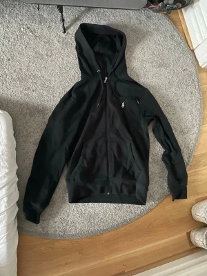 Svart hoodie från Polo Ralph Lauren - Snygg svart hoodie med dragkedja från Polo Ralph Lauren. Klassisk design med huva och fickor framtill samt den ikoniska loggan broderad på bröstet. Perfekt för en avslappnad och stilren look. Tillverkad i mjukt bomullsmaterial.