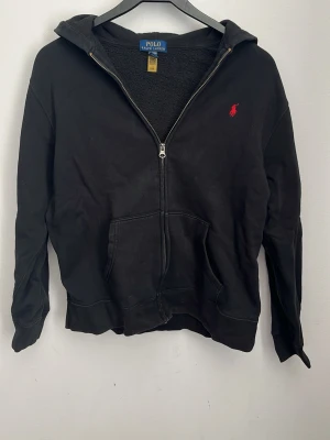 Svart hoodie från Polo Ralph Lauren - Svart hoodie med dragkedja från Polo Ralph Lauren. XL i barn storlek, S i vuxenstorlek. Klassisk design med huva, två fickor framtill och röd broderad logga på bröstet. Mjuk insida och ribbade muddar vid ärmslut och nederkant. Perfekt för en avslappnad och stilren look.