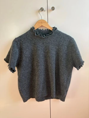 Grå stickad topp med volangkant - Mysig grå stickad topp med korta ärmar och volangkant vid halsringning och ärmslut. Tröjan har en rak passform och är tillverkad i ett mjukt material som känns skönt mot huden. Perfekt för lager på lager och enkel att styla. Onesize
