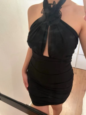 Svart kort halterneckklänning - Snygg svart kort klänning med halterneck och dekorativ rosett vid halsen. Klänningen har en liten slits framtill och är figurnära med draperade detaljer. Perfekt för dig som vill sticka ut med en elegant och trendig look. Prislapp är fof på!