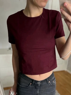 Vinröd croppad topp från H&M - Snygg vinröd croppad topp från H&M i mjuk bomull. T-shirten har rund halsringning och korta ärmar, perfekt till jeans eller kjol. Enkel och stilren design som är lätt att matcha.