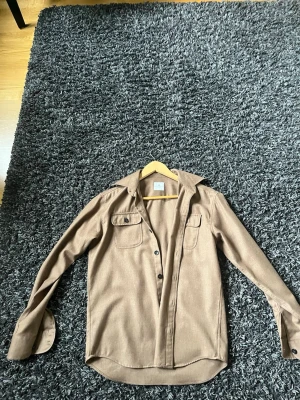 Beige overshirt från John Henric - Stilren beige overshirt från John Henric med klassisk krage och två bröstfickor med lock. Skjortan har knäppning framtill och är tillverkad i ett mjukt tyg som ger en avslappnad men snygg look. Perfekt att bära öppen över en t-shirt. Skick 10/10, så gott som ny.