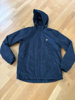 Svart vindjacka från Lyle & Scott - Snygg svart vindjacka från Lyle & Scott med huva och dragkedja framtill. Jackan har två fickor med knappar och det klassiska gula logobroderiet på bröstet. Lättviktigt material och meshfoder. Använd fåtal gånger. 