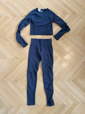 Mörkblått sömlöst träningsset - Säljer ett mörkblått sömlöst träningsset från lager 157 med långärmad crop top och höga tights. Setet är gjort i stretchigt material som sitter tajt och bekvämt, perfekt för gym eller yoga. Toppen har rund hals och tightsen har bred midjeresår för extra stöd.