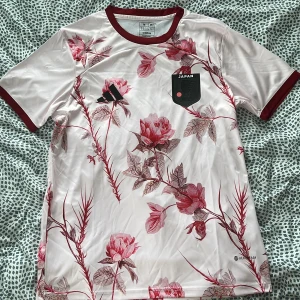 Japan fotbollströja med blommönster från Adidas - Säljer en Japan fotbollströja från Adidas i slim fit. Tröjan är vit med röda och rosa blommor, röda muddar och krage. Japan-emblem på bröstet och Adidas-logga. Perfekt för dig som gillar unik design! Har tag och original påsen som tröjan kom från. 