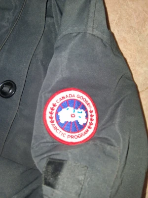 Svart parkajacka från Canada Goose - Svart parkajacka från Canada Goose med klassisk logotyp på ärmen. Jackan har huva, knäppning med stora knappar och är tillverkad i slitstarkt material. Perfekt för kalla dagar och har en snygg, figurnära passform.
