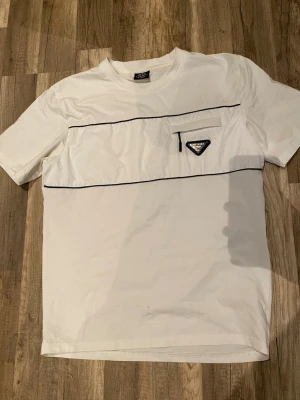 Vit t-shirt från Prada med ficka - Vit t-shirt från Prada med svarta detaljer och en liten ficka med dragkedja på bröstet. Klassisk passform och rund halsringning. Snyggt Prada-märke framtill och diskreta svarta linjer som ger en stilren look.