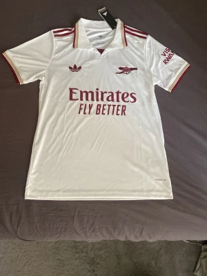 Vit Arsenal fotbollströja Adidas - Snygg vit Arsenal fotbollströja från Adidas med röda detaljer och klubbmärke på bröstet. Tröjan har korta ärmar, klassisk krage och röd text 'Emirates Fly Better' framtill. Tillverkad i lätt och ventilerande material, perfekt för träning eller match.