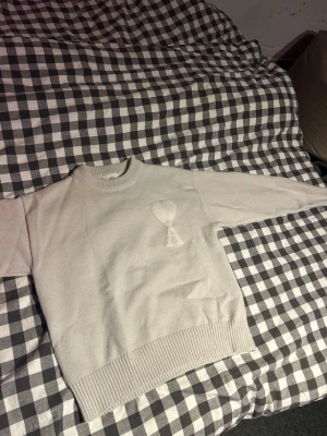 Beige stickad tröja från Ami Paris - Säljer en beige stickad tröja från Ami Paris med deras ikoniska hjärta och A-logga på bröstet. Tröjan har rund halsringning, ribbade muddar och är gjord i mjukt material. Perfekt för dig som gillar stilrena och trendiga plagg.