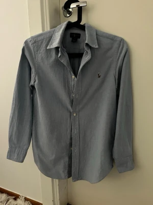 Blå skjorta från Ralph Lauren - Klassisk blå skjorta från Ralph Lauren med broderad logga på bröstet. Skjortan har lång ärm, knappar framtill och en stilren krage. Perfekt för en clean och tidlös look.