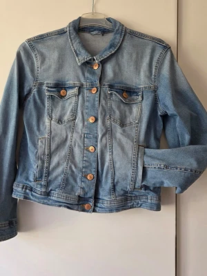 Klassisk ljusblå jeansjacka - Snygg ljusblå jeansjacka med klassisk passform och bruna knappar framtill. Jackan har två bröstfickor med lock och knappar samt två sidofickor. Perfekt för dig som vill ha en tidlös och trendig look.