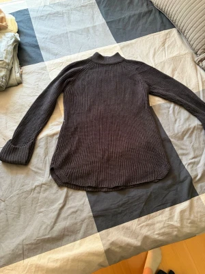 Mörkgrå stickad polotröja - Mörkgrå stickad polotröja med lång ärm och ribbstickad struktur. Tröjan har en hög krage och rundad nederkant, vilket ger en avslappnad och snygg passform. Perfekt för lager-på-lager och enkel att matcha med jeans eller kjol.