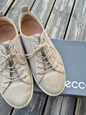 Beige loafers från Ecco i skinn - Snygga beige loafers från Ecco i mjukt skinn med perforerade detaljer på sidorna och klassisk snörning. Skorna har en låg, vit sula och en stilren, minimalistisk design som passar till många outfits. Perfekta för dig som gillar bekväma och stilrena skor.