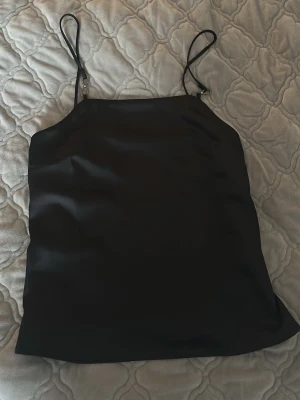 Svart satinlinnetopp från H&M - Säljer en svart linnetopp från H&M i glansigt satinmaterial. Toppen har tunna justerbara axelband och en enkel, stilren design som passar perfekt till många olika looks.pris kan diskuteras och den e använd 1 gång 🤗