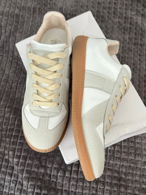 Vita sneakers med beige detaljer - Stilrena sneakers i vitt läder med beige mockadetaljer och beige snörning. Yttersulan är i gummi med en klassisk brun färg. Snygg och tidlös design som passar till många outfits.