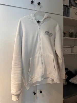Vit hoodie med tryck från Night Sky - Vit hoodie med dragkedja från Night Sky. Stor svartvit ving- och textprint på ryggen samt mindre logga på bröstet. Mjuk bomullskänsla, huva och två stora fickor framtill. Perfekt för en chill och stilren look.