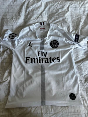 Vit PSG matchtröja Jordan x Fly Emirates - Säljer en vit PSG matchtröja i samarbete med Jordan och Fly Emirates. Tröjan har svarta detaljer, PSG-logga på bröstet, Champions League-märke på ärmen och diskret rand i mitten. Tillverkad i lätt och ventilerande material, perfekt för fotboll eller träning.