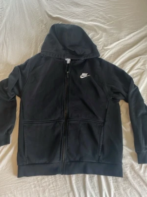 Svart Nike hoodie med dragkedja - Ny Svart hoodie från Nike med vit broderad logga på bröstet. Tröjan har dragkedja framtill, två stora fickor och en klassisk huva. Perfekt för en avslappnad och sportig stil. Tillverkad i mjukt material för extra komfort.