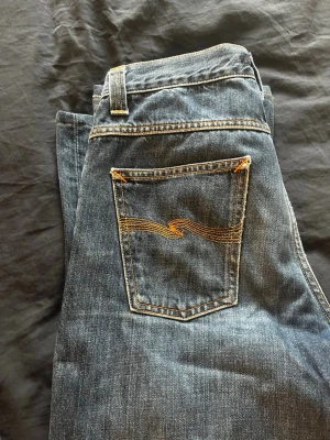 Blå jeans från Nudie Jeans 30/32 - Snygga blå jeans från Nudie Jeans med klassisk femficksdesign och gul kontrastsöm på bakfickan. Jeansen har en rak passform och är tillverkade i slitstarkt denim. Perfekta för en avslappnad och trendig stil.