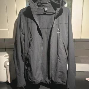 Svart vindjacka från Haglöfs med huva - Svart vindjacka från Haglöfs med minimalistisk design, hel dragkedja och två snedställda fickor med dragkedja. Jackan har huva och diskret logga på bröstet. Tillverkad i slitstark polyester, perfekt för blåsiga dagar.