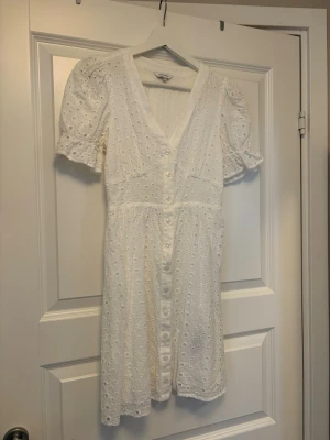 Vit klänning - Superfin vit kortklänning med broderade detaljer och v-ringning. Klänningen har korta puffärmar med volangkant och knappar framtill. Perfekt för sommarens alla tillfällen!