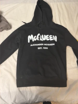 Svart hoodie från Alexander McQueen - Cool svart hoodie från Alexander McQueen med vit logotyp och text på bröstet. Klassisk huva med dragsko och stor magficka framtill. Materialet är mjukt och bekvämt, perfekt för att chilla eller hänga med kompisar. Snygg och stilren design med tydlig branding.