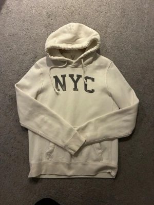 Vit hoodie med NYC-tryck Abercrombie - Vit hoodie från Abercrombie & Fitch med grått NYC-tryck på bröstet. Klassisk huva med dragsko och känguruficka framtill. Mjuk bomullskänsla och ribbade muddar vid ärmslut och nederkant. Perfekt för en avslappnad streetstyle-look.