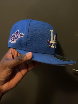 Blå LA Dodgers keps med brodyr - Snygg blå LA Dodgers keps från New Era med broderad logga och blommotiv framtill. Kepsen har platt skärm, lila undersida och World Series-märke på sidan. Tillverkad i bomull och polyester, perfekt för en sportig och trendig look.
