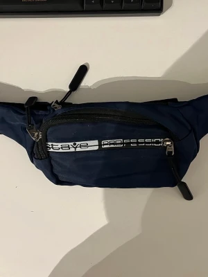 Axelväska Stay Professional - Snygg marinblå magväska från Stay Professional med svart justerbart band och dragkedja. Väskan har ett vitt tryck framtill och flera fack med dragkedja, perfekt för att bära med sig det viktigaste. Materialet är slitstark textil.