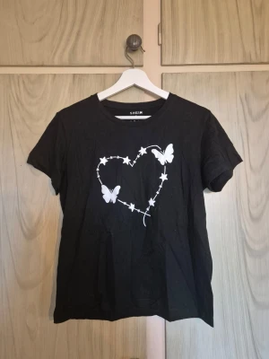 Svart t-shirt med hjärta och fjärilar - Svart t-shirt med vitt tryck av ett hjärta format av stjärnor och två fjärilar på bröstet. Klassisk rund hals och korta ärmar. Perfekt för en avslappnad och trendig look.     Alla kläder rollas innan dom skickas hem då det finns katt i hemmet🐈 Jag stryker och tvättar INTE innan jag skickar det får du stå för själv så det blir som du vill ha det🤗