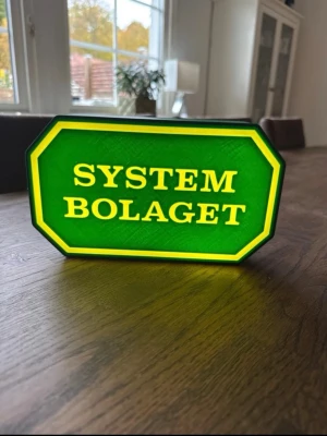 Systembolaget skylt med LED-belysning - Grön och gul skylt med texten 'Systembolaget', inspirerad av butikens klassiska logga. Skylten är tillverkad i plast och har inbyggd LED-belysning som lyser upp texten. Perfekt som dekoration till hemmabaren eller som rolig inredningsdetalj.