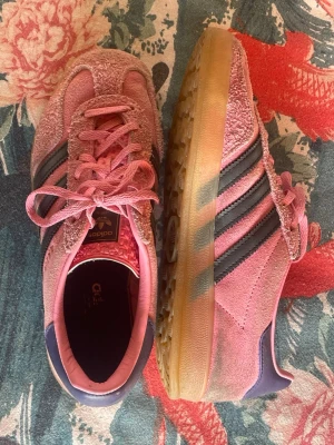 Adidas Gazelle rosa - Säljer ett par rosa Adidas Gazelle sneakers i mocka med klassiska svarta ränder på sidorna och gummisula. Skorna har rosa snören och detaljer i mörkblått vid hälen. Perfekta för dig som vill sticka ut med färg och ikonisk retrostil.   !!endast använt ett fåtal gånger!!