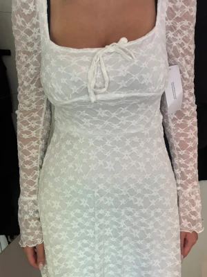 Vit spetsklänning med långa ärmar - Superfin vit spetsklänning från NA-KD med långa ärmar och knyt framtill. Klänningen har blommigt spetsmönster, figurnära passform och är kort i modellen. Perfekt för sommarens alla tillfällen!