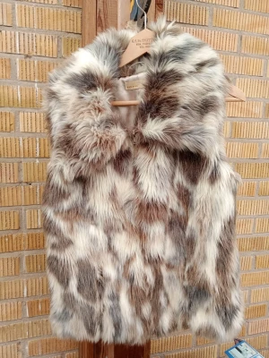 Fluffig flerfärgad pälsväst - Snygg och trendig pälsväst i fluffigt fuskpälsmaterial med mixade nyanser av brunt, grått, vitt och beige. Västen har en klassisk krage och är perfekt för att ge din outfit en extra edge. Stängs framtill och har en mjuk insida.
