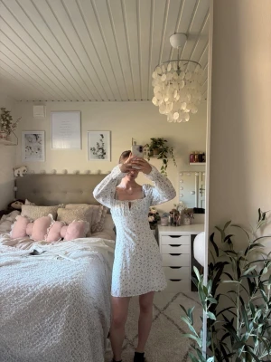 Vit blommig kortklänning med puffärm - Supersöt vit kortklänning med små blommor och puffiga långärmar. Klänningen har en fyrkantig ringning, liten nyckelhålsdetalj framtill och sitter figurnära upptill. Perfekt för sommarens alla tillfällen!