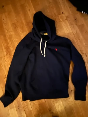 Mörkblå hoodie från Polo Ralph Lauren - Mörkblå hoodie från Polo Ralph Lauren med vit snörning i huvan och röd broderad logga på bröstet. Klassisk känguruficka framtill och mjukt material som känns skönt mot huden. Perfekt för dig som gillar stilren och enkel streetwear. Perfekt skick  inge hål, märken eller dålig lukt, passar L ganska bra fast den är M. Kvitto finns! Skriv vid funderingar 