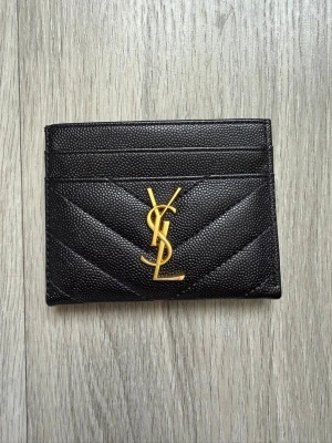 Svart Saint Laurent kortfodral - Snyggt svart kortfodral från Saint Laurent i präglat läder med guldigt YSL-logo framtill. Fodralet har flera kortfack och stilren design med diskreta sömmar i chevronmönster. Perfekt för dig som vill ha en lyxig och smidig accessoar.