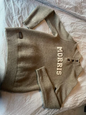 Grå stickad tröja från Morris - Snygg grå stickad tröja från Morris med broderad logga framtill. Tröjan har ribbade muddar, hög krage och tre knappar vid halsen. Perfekt för dig som gillar klassisk och stilren design.