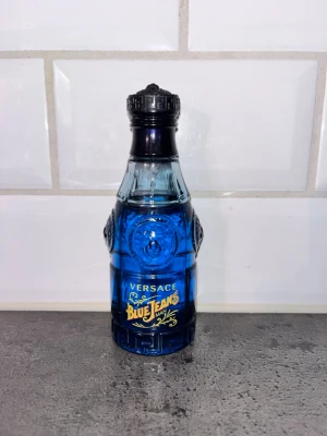 Versace Blue Jeans Man parfym - Versace Blue Jeans Man parfym i en cool blå glasflaska med detaljerad design och gul text. Flaskan har en unik form och är fylld med blått innehåll. Perfekt för dig som gillar klassiska och fräscha dofter. Använd ett par få gånger, Utan Låda. 75ml orginalt men har användt den ett par få gånger så skulle säga 70ml