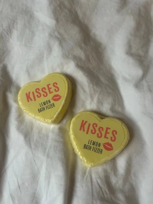 Kisses Lemon Bath Fizzer hjärtformad - Två stycken hjärtformade badbomber med citrondoft, inslagna i gul plast med texten 'Kisses Lemon Bath Fizzer' och en röd kyssmun. Perfekt för ett lyxigt bad och en fräsch känsla. Varje badbomb väger 60 g.