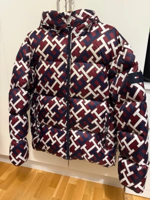 Tommy Hilfiger pufferjacka mönstrad - Säljer en fet pufferjacka från Tommy Hilfiger med vinrött, vitt och marinblått TH-mönster över hela jackan. Jackan har huva, dragkedja framtill och logga på ärmen. Perfekt för dig som vill sticka ut i vinterkylan.